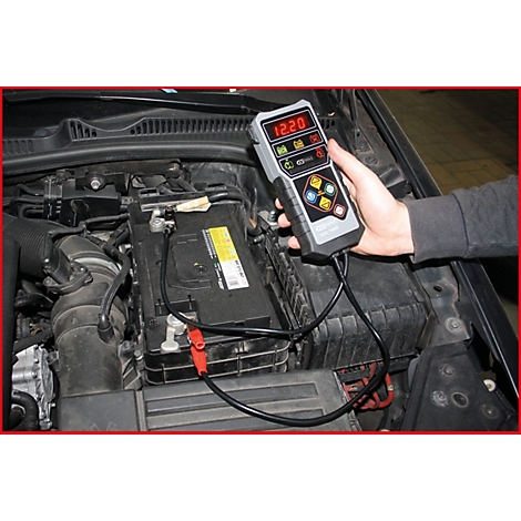 Person misst mit einem Batterietester die Spannung einer Autobatterie im Motorraum. Das Messgerät zeigt 12,20 Volt an.